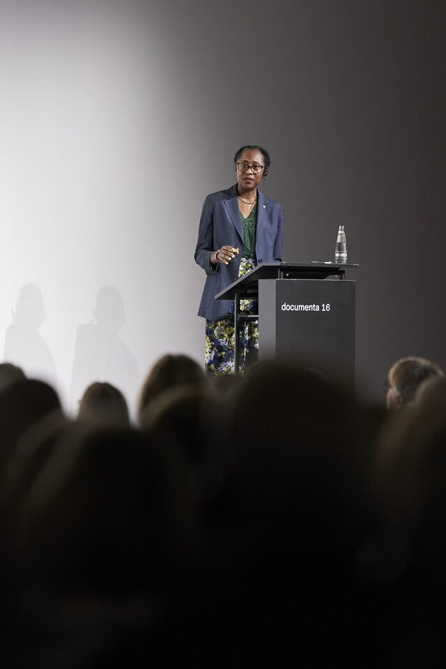 Naomi Beckwith is seen on a stage, giving a lecture in front of numerous people
